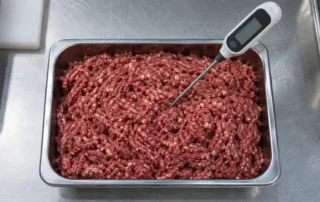 Bandeja de carne picada de vacuno cruda sobre superficie fría, con termómetro de sonda insertado midiendo la temperatura del producto